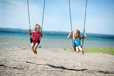 Two girls on swings with sand on the ground and water in the background