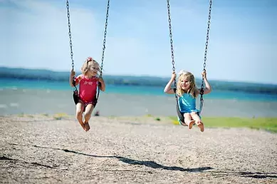 Two girls on swings with sand on the ground and water in the background